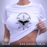Футболка дух предків, біла, складена на столі, фон - світло-сірий, принт чорного кольору з птахом та декоративними елементами, на комірці видна етикетка.