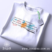 Футболка душа співає, біла, на столі, з верхнього ракурсу, фон світло-сірий, принт з текстом у різних кольорах: червоний, зелений, чорний, з елементами графіки, з етикеткою на комірці.