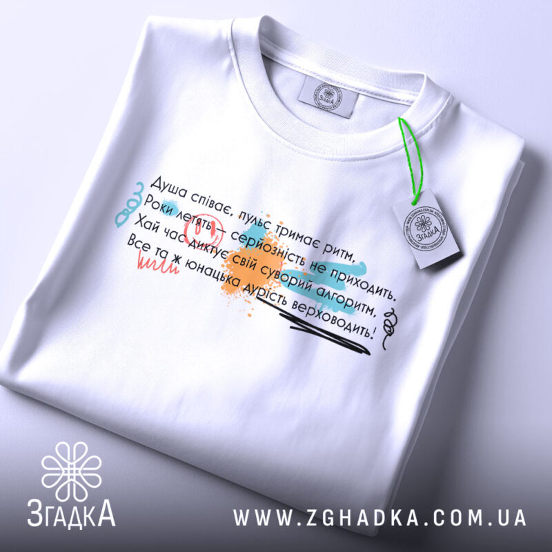 Футболка душа співає, біла, на столі, з верхнього ракурсу, фон світло-сірий, принт з текстом у різних кольорах: червоний, зелений, чорний, з елементами графіки, з етикеткою на комірці.