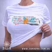 Футболка душа співає, біла, на столі, з верхнього ракурсу, фон світло-сірий, принт з текстом у різних кольорах: червоний, зелений, чорний, з елементами графіки, з етикеткою на комірці.