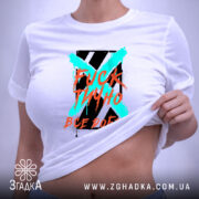 Футболка фактично все добре. Біла футболка, складена на поверхні, з принтом у центрі. Принт містить текст і кольорові елементи, а фон світлий і нейтральний.