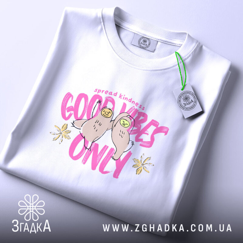 Футболка Good Vibes Only білого кольору, складена на поверхні, з видимим принтом, що містить малюнок двох сов та напис 'Good Vibes Only' в рожевих тонах. На футболці також видно етикетку з зеленим шнурком.