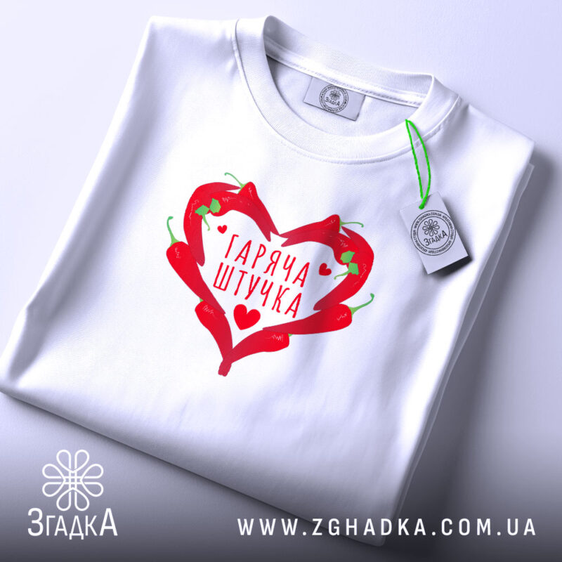 Футболка гаряча штучка, біла, складена на поверхні, з принтом у вигляді червоного серця з перцями, фон світло-сірий, етикетка з зеленим шнурком додає акцент.
