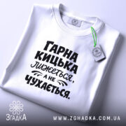 Футболка гарна кицька лижеться. Біла футболка, складена на поверхні, з чорним принтом. Фон світло-сірий, на футболці видно текст: 'ГАРНА КИЦЬКА ЛИЖЕТЬСЯ, А НЕ ЧУХАЄТЬСЯ', а також етикетка з зеленим шнурком.