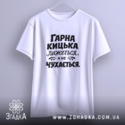 Футболка гарна кицька лижеться. Біла футболка, підвішена на вішалці, з чорним принтом. Фон світло-сірий, текст на футболці: 'ГАРНА КИЦЬКА ЛИЖЕТЬСЯ, А НЕ ЧУХАЄТЬСЯ'.