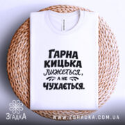 Футболка гарна кицька лижеться. Біла футболка, складена на плетеній основі, з чорним принтом. Фон світло-сірий, текст на футболці: 'ГАРНА КИЦЬКА ЛИЖЕТЬСЯ, А НЕ ЧУХАЄТЬСЯ'.