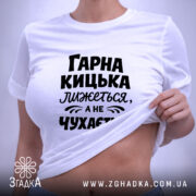 Футболка гарна кицька лижеться. Біла футболка, складена на поверхні, з чорним принтом. Фон світло-сірий, на футболці видно текст: 'ГАРНА КИЦЬКА ЛИЖЕТЬСЯ, А НЕ ЧУХАЄТЬСЯ', а також етикетка з зеленим шнурком.