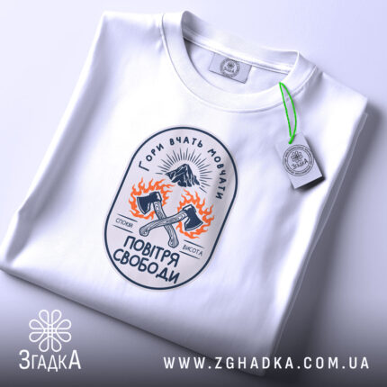 Футболка гори вчать мовчати, біла, зображена зверху, фон світло-фіолетовий, принт з малюнком двох сокир, оточених вогнем, та написом 'Гори вчать мовчати' у верхній частині, на комірці є етикетка.