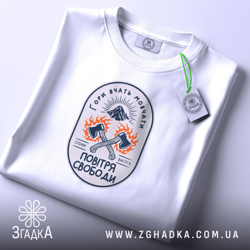 Футболка гори вчать мовчати, біла, зображена зверху, фон світло-фіолетовий, принт з малюнком двох сокир, оточених вогнем, та написом 'Гори вчать мовчати' у верхній частині, на комірці є етикетка.