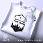 Футболка гуцул не нервує, біла, на столі, з коротким рукавом, фон світло-сірий, принт чорний з написом "Гуцул не нервує – Гуцул планує" та зображенням лісу, з етикеткою з зеленим шнурком.