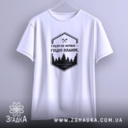 Футболка гуцул не нервує, біла, підвішена на вішалці, фон світло-сірий, принт чорний з написом "Гуцул не нервує – Гуцул планує" та зображенням лісу.