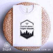 Футболка гуцул не нервує, біла, на плетеній кошику, фон світло-сірий, принт чорний з написом "Гуцул не нервує – Гуцул планує" та зображенням лісу.