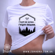 Футболка гуцул не нервує, біла, на столі, з коротким рукавом, фон світло-сірий, принт чорний з написом "Гуцул не нервує – Гуцул планує" та зображенням лісу, з етикеткою з зеленим шнурком.