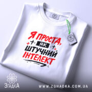 Футболка я як штучний інтелект, біла, зображена в складеному вигляді, фон світло-сірий, принт червоними і чорними літерами з написом 'Я ПРОСТА, ЯК ШТУЧНИЙ ІНТЕЛЕКТ', з етикеткою на зеленій нитці.