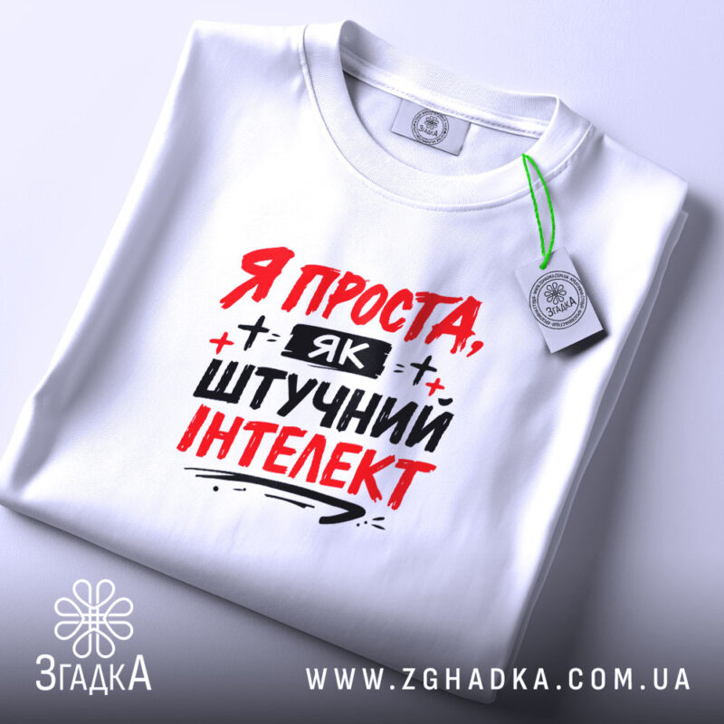 Футболка я як штучний інтелект, біла, зображена в складеному вигляді, фон світло-сірий, принт червоними і чорними літерами з написом 'Я ПРОСТА, ЯК ШТУЧНИЙ ІНТЕЛЕКТ', з етикеткою на зеленій нитці.