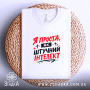 Футболка я як штучний інтелект, біла, зображена в складеному вигляді на плетеній основі, фон світло-сірий, принт червоними і чорними літерами з написом 'Я ПРОСТА, ЯК ШТУЧНИЙ ІНТЕЛЕКТ'.
