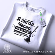 Футболка я янгол, білого кольору, складена на поверхні, фон світло-сірий, принт чорного кольору з текстом 'Я янгол. Чесно. Але на мітлі - швидше.' та малюнком мітли, з етикеткою зеленого кольору.