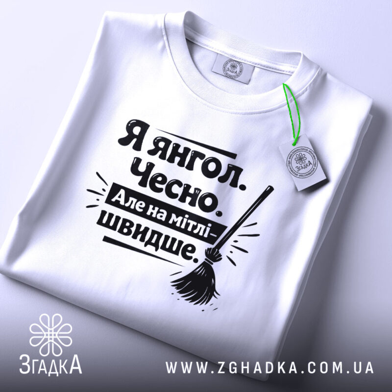 Футболка я янгол, білого кольору, складена на поверхні, фон світло-сірий, принт чорного кольору з текстом 'Я янгол. Чесно. Але на мітлі - швидше.' та малюнком мітли, з етикеткою зеленого кольору.