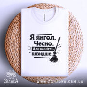 Футболка я янгол, білого кольору, складена на плетеній кошику, фон білий, принт чорного кольору з текстом 'Я янгол. Чесно. Але на мітлі - швидше.' та малюнком мітли.