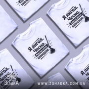 Футболка я янгол, білого кольору, зображена на моделі з коротким рукавом, фон світло-фіолетовий, принт чорного кольору з текстом 'Я янгол. Чесно. Але на мітлі - швидше.' та малюнком мітли.