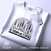 Футболка я обираю гори, біла, складена на поверхні, з принтом у вигляді лісу та місяця, фон світло-сірий, на етикетці зелена стрічка.