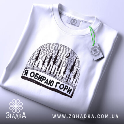 Футболка я обираю гори, біла, складена на поверхні, з принтом у вигляді лісу та місяця, фон світло-сірий, на етикетці зелена стрічка.