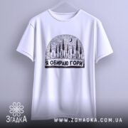 Футболка я обираю гори, біла, підвішена на вішалці, з принтом у вигляді лісу та місяця, фон світло-сірий, деталі принта чіткі та контрастні.