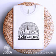 Футболка я обираю гори, біла, складена на плетеній основі, з принтом у вигляді лісу та місяця, фон світло-сірий, деталі принта чіткі та контрастні.