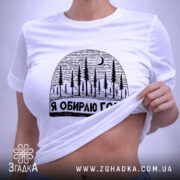 Футболка я обираю гори, біла, складена на поверхні, з принтом у вигляді лісу та місяця, фон світло-сірий, на етикетці зелена стрічка.