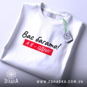 Футболка я одна, білого кольору, складена на поверхні, фон світло-сірий, принт чорного кольору з червоним фоном, текст "Вас багато! А я – одна!", на комірці є етикетка.