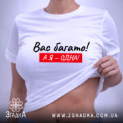 Футболка я одна, білого кольору, складена на поверхні, фон світло-сірий, принт чорного кольору з червоним фоном, текст "Вас багато! А я – одна!", на комірці є етикетка.