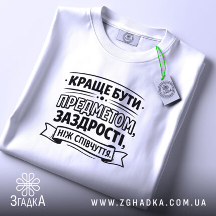 Футболка я предмет заздрості, біла, лежить на рівній поверхні, ракурс зверху, принт чорного кольору з текстом, прикріплений етикеткою зеленого кольору.