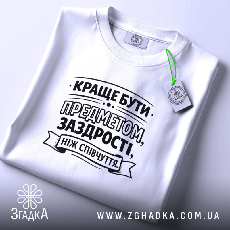 Футболка я предмет заздрості, біла, лежить на рівній поверхні, ракурс зверху, принт чорного кольору з текстом, прикріплений етикеткою зеленого кольору.