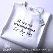 Футболка я проста як квантова фізика. Біла футболка, складена на поверхні, з ракурсом зверху. Фон світлий, на футболці чорний принт з текстом і малюнками, а також зелена етикетка.