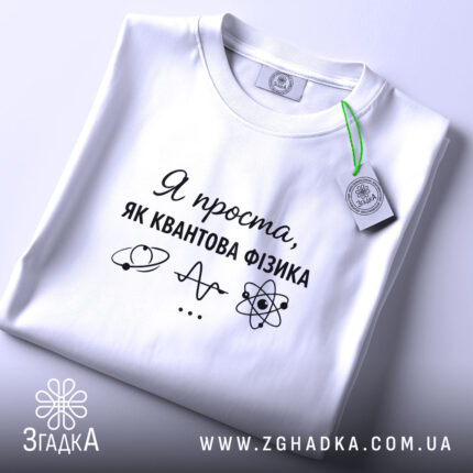 Футболка я проста як квантова фізика. Біла футболка, складена на поверхні, з ракурсом зверху. Фон світлий, на футболці чорний принт з текстом і малюнками, а також зелена етикетка.