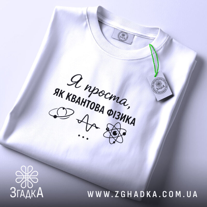 Футболка я проста як квантова фізика. Біла футболка, складена на поверхні, з ракурсом зверху. Фон світлий, на футболці чорний принт з текстом і малюнками, а також зелена етикетка.