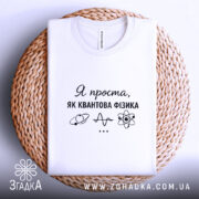 Футболка я проста як квантова фізика. Біла футболка, складена на плетеній основі, з ракурсом зверху. Фон світлий, на футболці чорний принт з текстом і малюнками, що символізують науку.