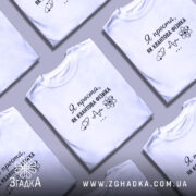 Футболка я проста як квантова фізика. Біла футболка, зображена на моделі, яка стоїть в ракурсі спереду. Фон ніжно-фіолетовий, на футболці чорний принт з текстом і малюнками, що символізують науку.