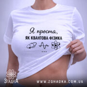 Футболка я проста як квантова фізика. Біла футболка, складена на поверхні, з ракурсом зверху. Фон світлий, на футболці чорний принт з текстом і малюнками, а також зелена етикетка.