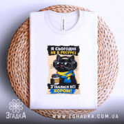 Футболка я сьогодні не в ресурсі. Біла футболка, розкладена на плетеній кошику, з принтом чорного кота в окулярах та навушниках, який тримає чашку кави. Фон світлий, деталі принта яскраві, текст українською мовою.