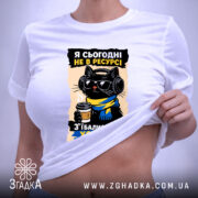 Футболка я сьогодні не в ресурсі. Біла футболка, розкладена на поверхні, з принтом чорного кота в окулярах та навушниках, який тримає чашку кави. Фон світло-фіолетовий, на футболці є етикетка з зеленим шнурком.