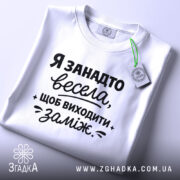 Футболка я занадто весела. Біла футболка, зображена зверху на світлому фоні. Принт чорного кольору з текстом, що говорить про веселий настрій, займає центральну частину. На комірці видно етикетку.