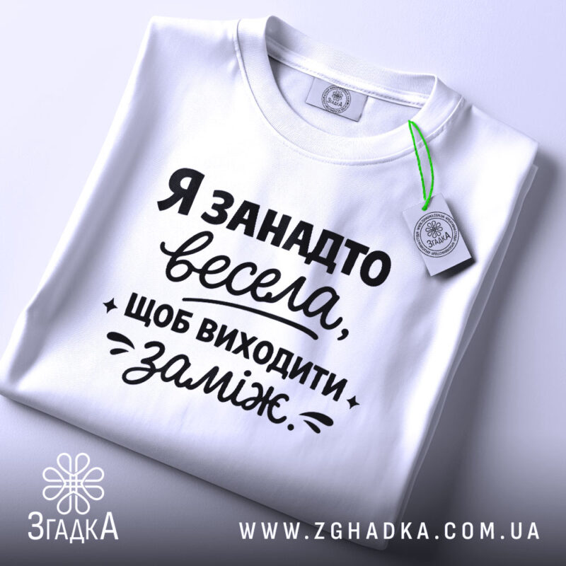Футболка я занадто весела. Біла футболка, зображена зверху на світлому фоні. Принт чорного кольору з текстом, що говорить про веселий настрій, займає центральну частину. На комірці видно етикетку.