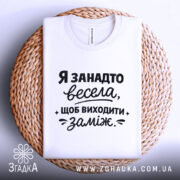 Футболка я занадто весела. Біла футболка, складена на плетеній підставці, зображена на світлому фоні. Принт чорного кольору з текстом, що говорить про веселий настрій, займає центральну частину.