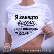 Футболка я занадто весела. Біла футболка, зображена зверху на світлому фоні. Принт чорного кольору з текстом, що говорить про веселий настрій, займає центральну частину. На комірці видно етикетку.