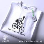 Футболка їду туди де добре, біла футболка, складена на поверхні, з принтом чорного кольору, на якому зображена лелека на велосипеді, фон світло-сірий, деталі принта яскраві та виразні.