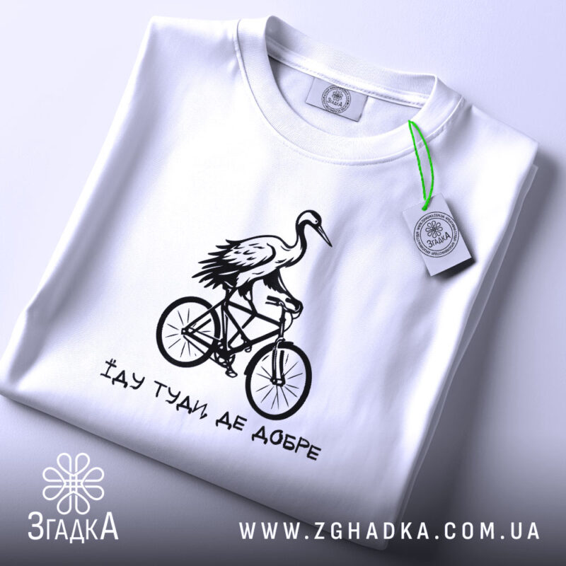 Футболка їду туди де добре, біла футболка, складена на поверхні, з принтом чорного кольору, на якому зображена лелека на велосипеді, фон світло-сірий, деталі принта яскраві та виразні.