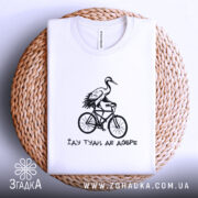 Футболка їду туди де добре, біла футболка, складена на плетеній кошику, з принтом чорного кольору, на якому зображена лелека на велосипеді, фон світло-блакитний, деталі принта яскраві та виразні.