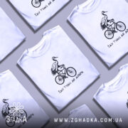 Футболка їду туди де добре, біла футболка на чоловікові, з принтом чорного кольору, на якому зображена лелека на велосипеді, фон світло-фіолетовий, деталі принта чіткі та контрастні.