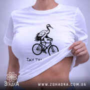 Футболка їду туди де добре, біла футболка, складена на поверхні, з принтом чорного кольору, на якому зображена лелека на велосипеді, фон світло-сірий, деталі принта яскраві та виразні.